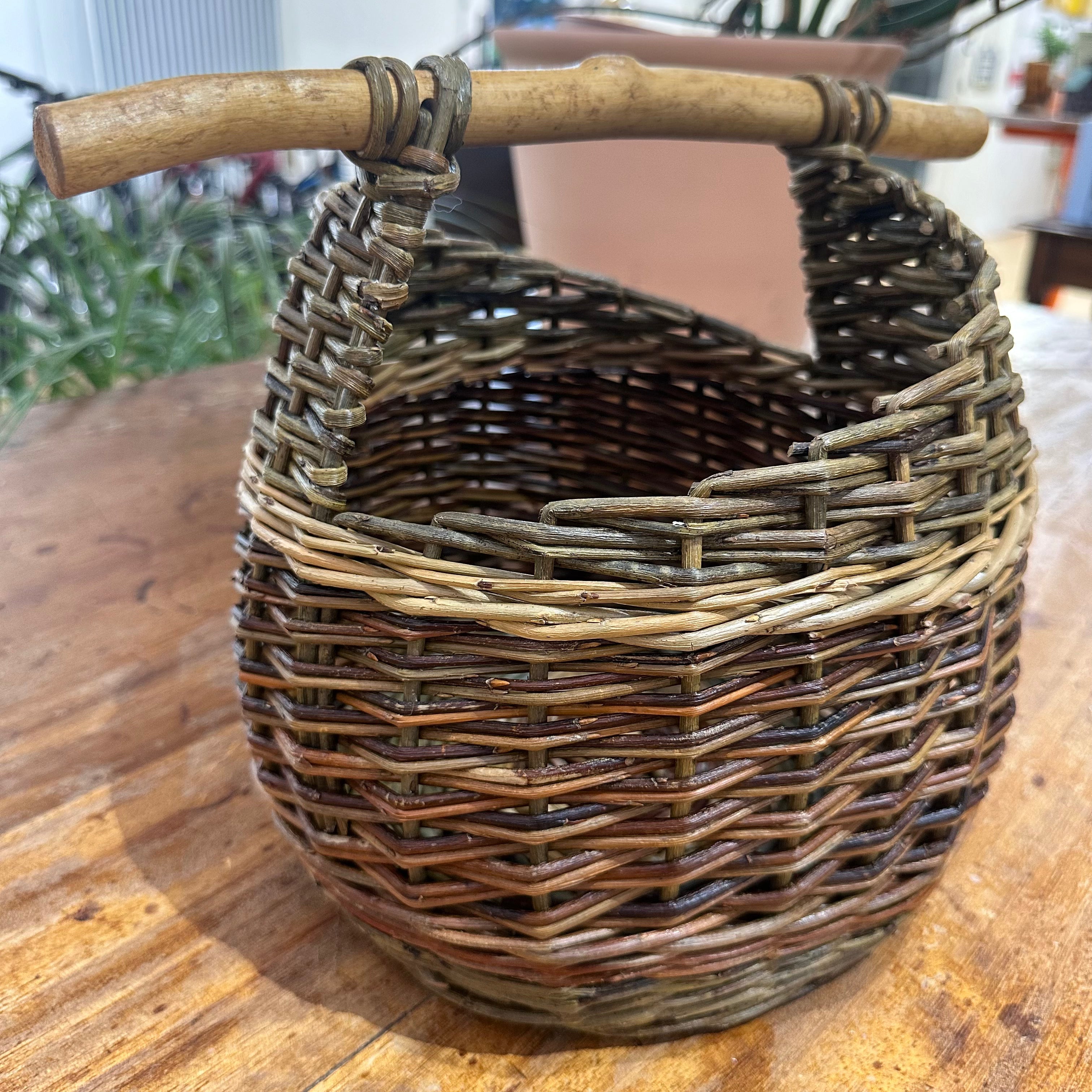 A large asymmetric basket made from brown willow with a handle, displayed on a wooden surface.