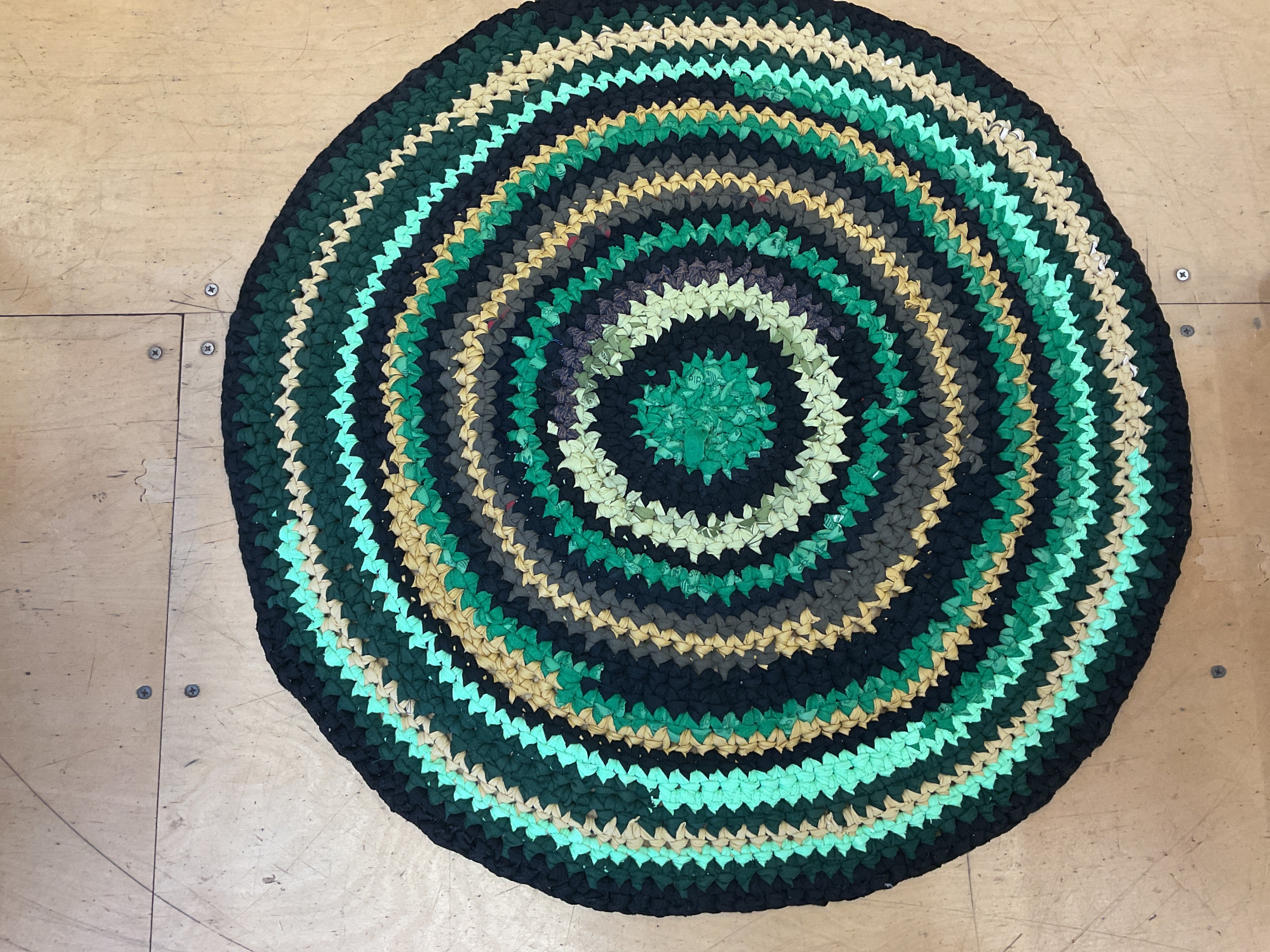 Round crochet rug made from upcycled t-shirts, with concentric circles of green, black, blue and beige on a wooden floor.