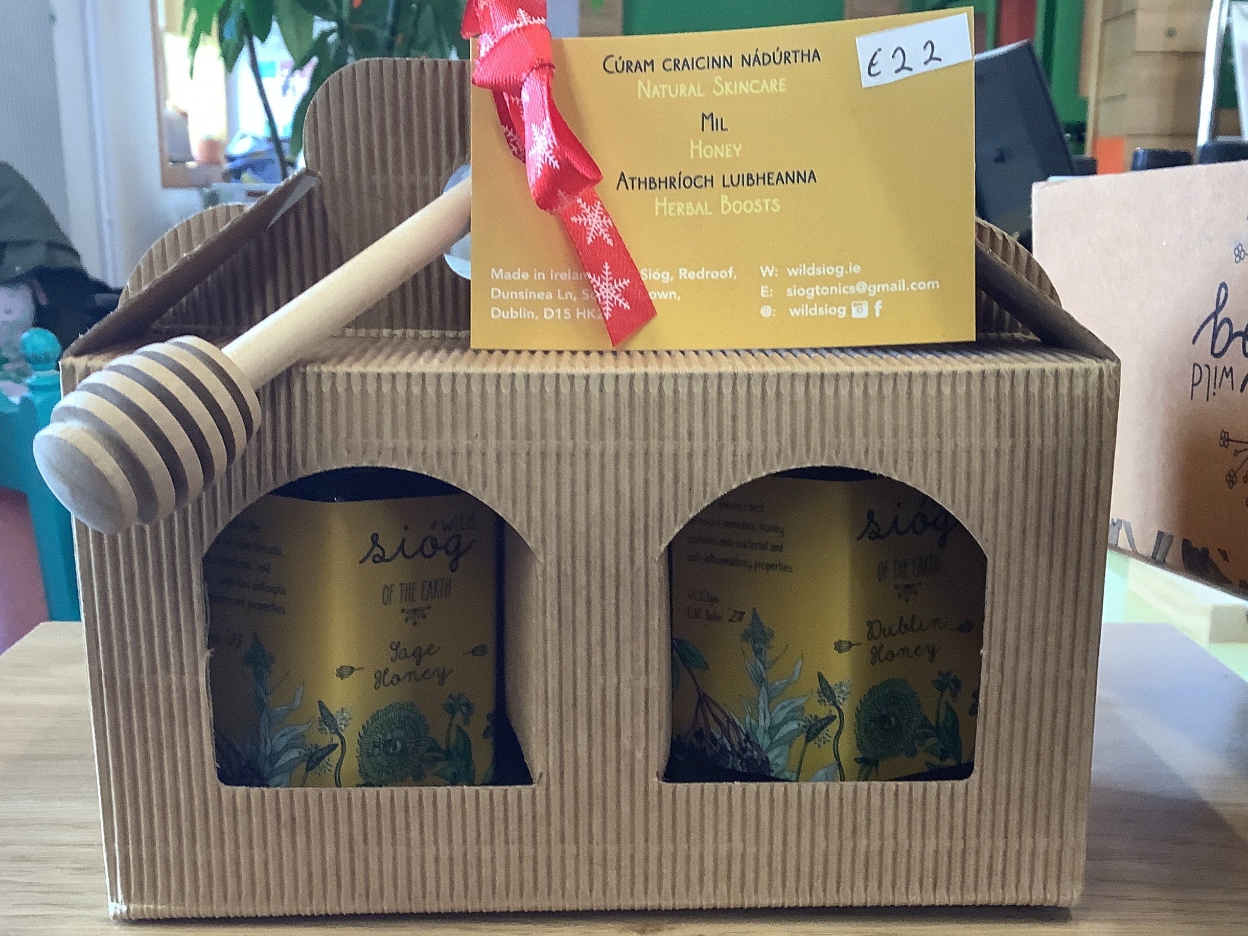 Two honey jars in a cardboard box with a honey dipper and a yellow label on a table.