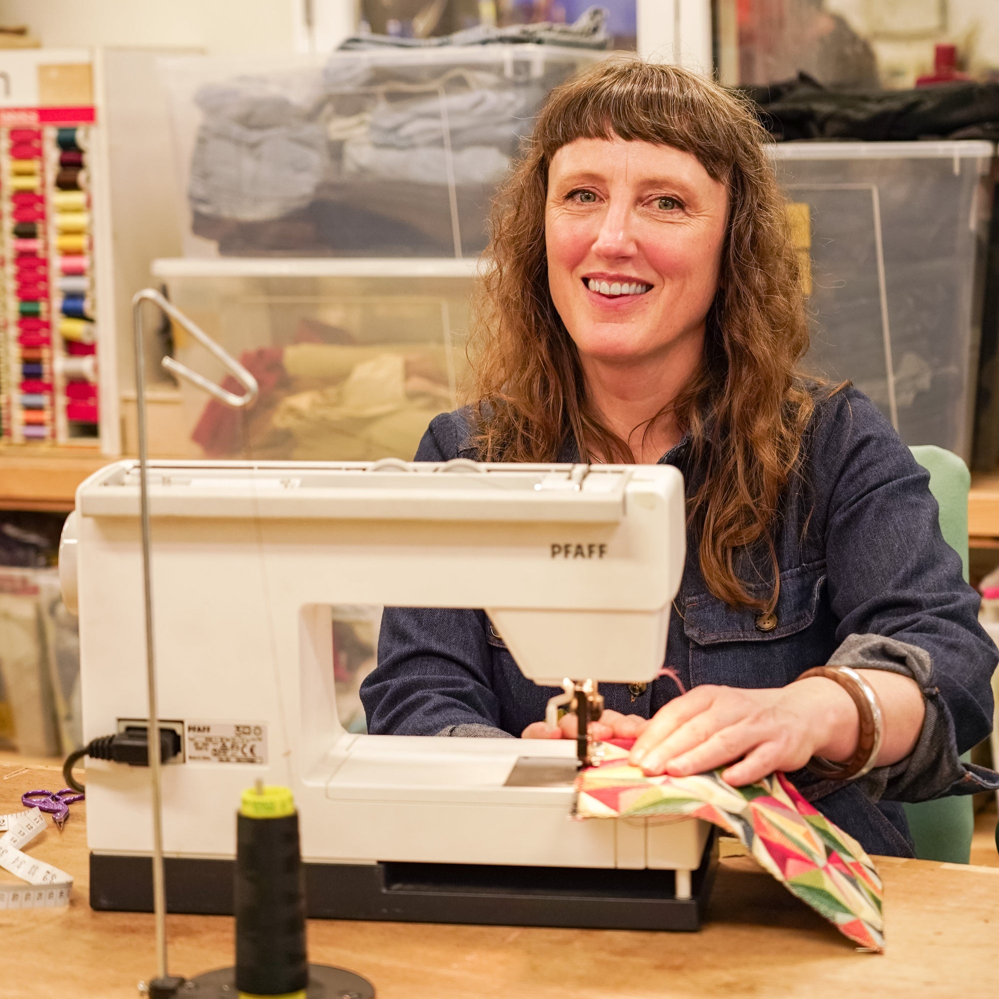 A woman smiling while operating a white Piastri sewing machine in a sewing room, with a spool of black thread on a stand in the foreground.