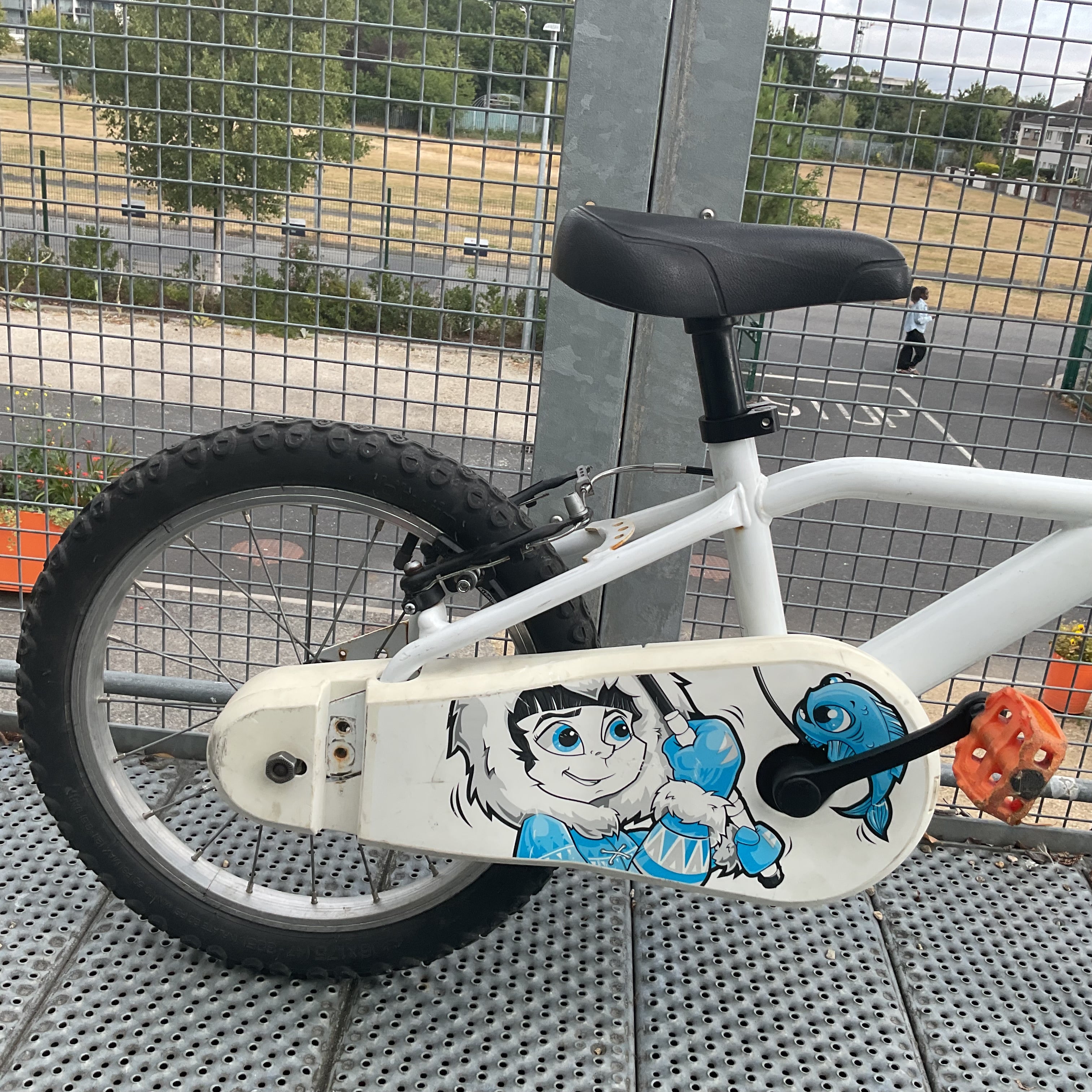 A children's bicycle with a white frame and black tires, featuring a custom hand-painted cartoon monkey and blue bird graphic on the chain guard. The bike is photographed outdoors on a metal grate floor in front of a wire fence.