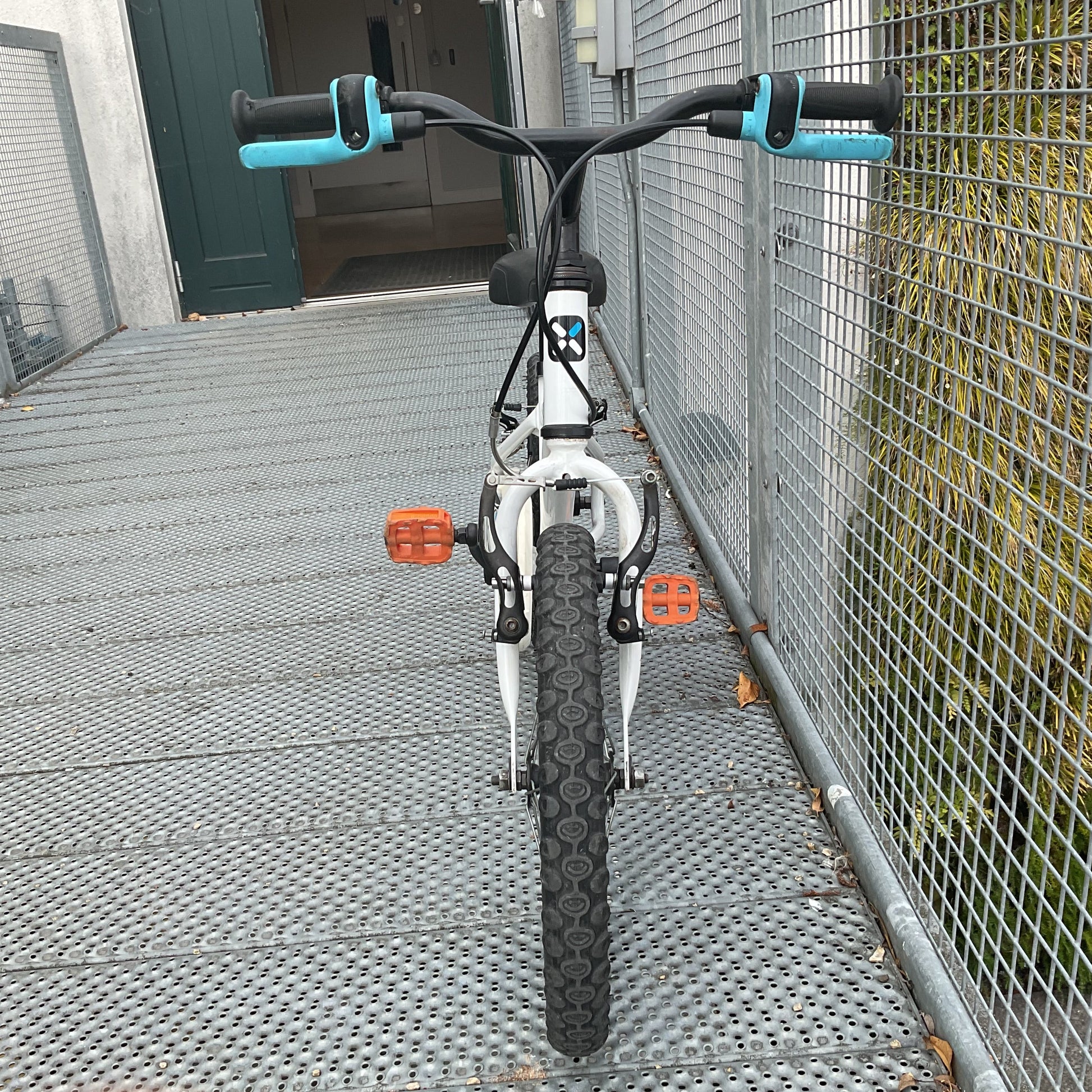 A wchildren's bicycle with a white frame, black tires, and turquoise handlebar grips, photographed from the front on a metal grate walkway next to a wire fence.