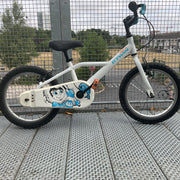 A wchildren's bicycle with a white frame, black tires, and turquoise handlebar grips, photographed from the front on a metal grate walkway next to a wire fence.