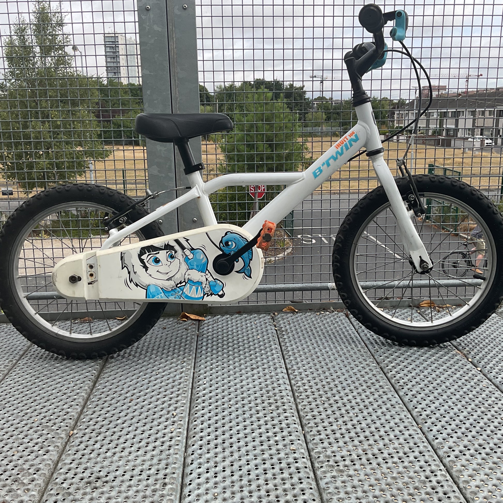 A wchildren's bicycle with a white frame, black tires, and turquoise handlebar grips, photographed from the front on a metal grate walkway next to a wire fence.