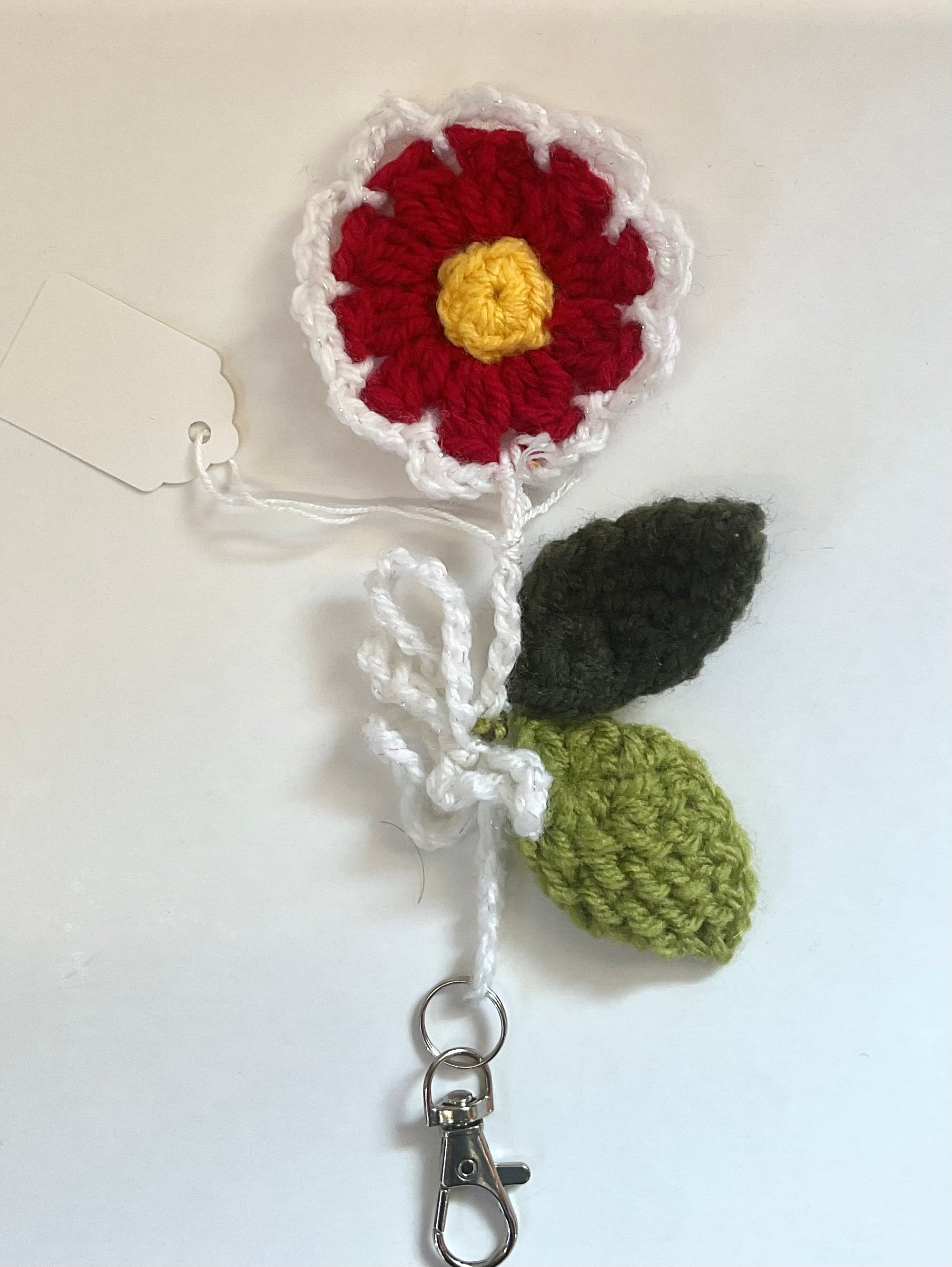 Crocheted flower keychain with red petals, yellow centre, green leaves, and  a white stem and white external petals and on a light background