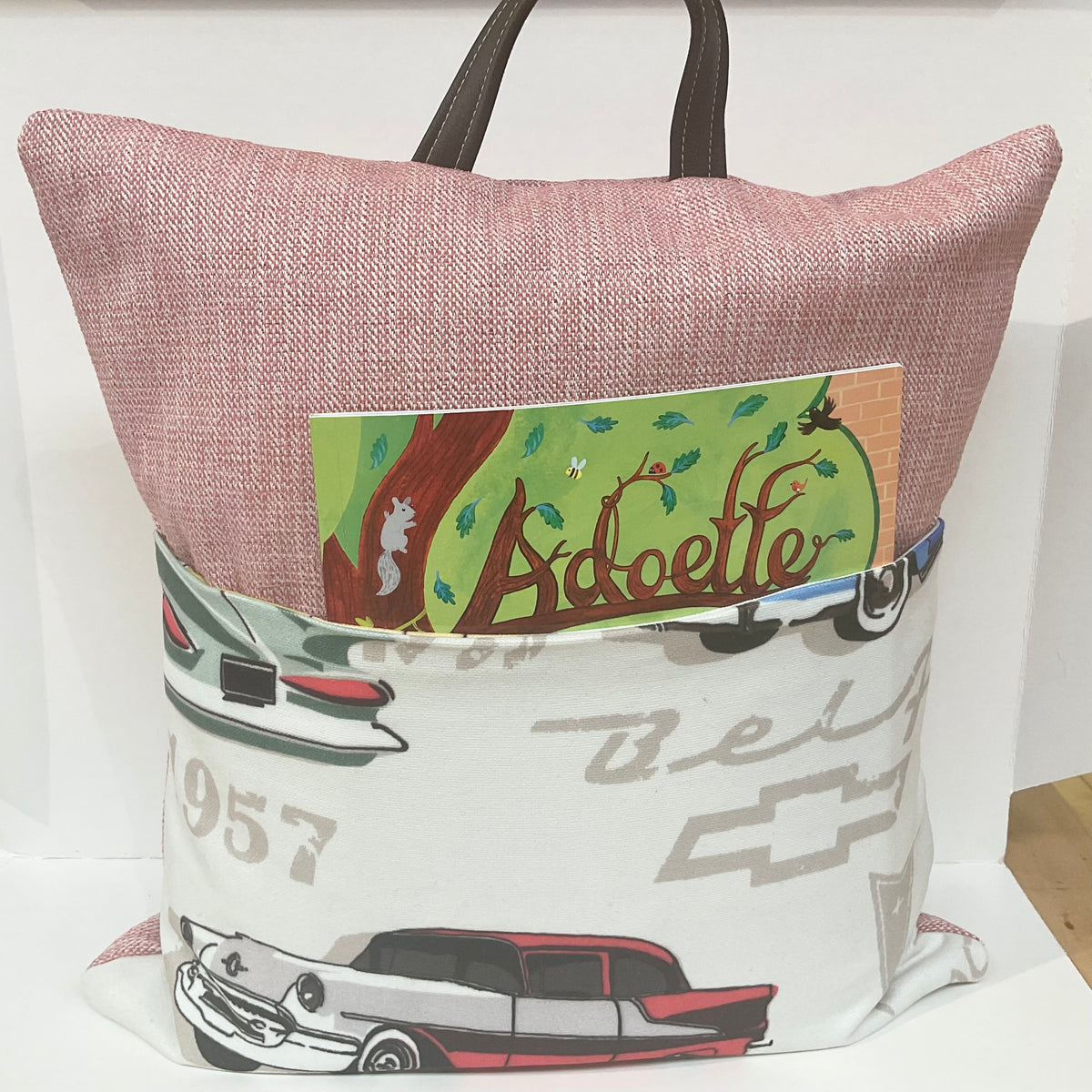 A fabric tote bag with a dusty pink woven upper section and dark brown handles, featuring a front pocket printed with vintage cars and the word 'Adoette' on a green card tucked into the pocket.