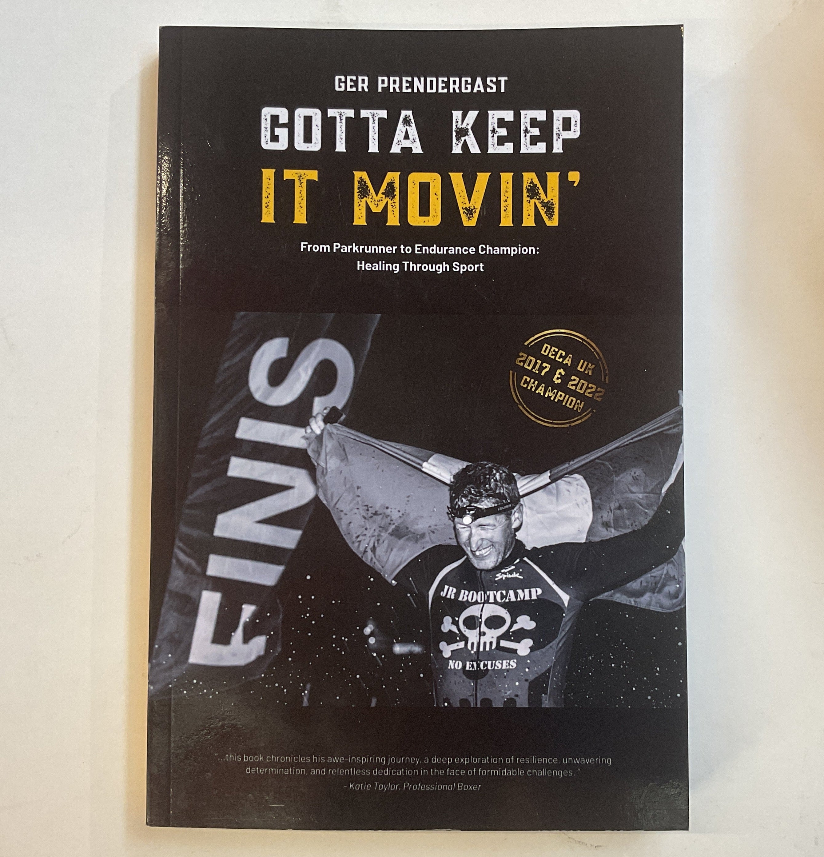 Book cover with text 'Gotta Keep It Movin'' and featuring athlete Ger Prendergast holding an Irish flag at the finish line of a race