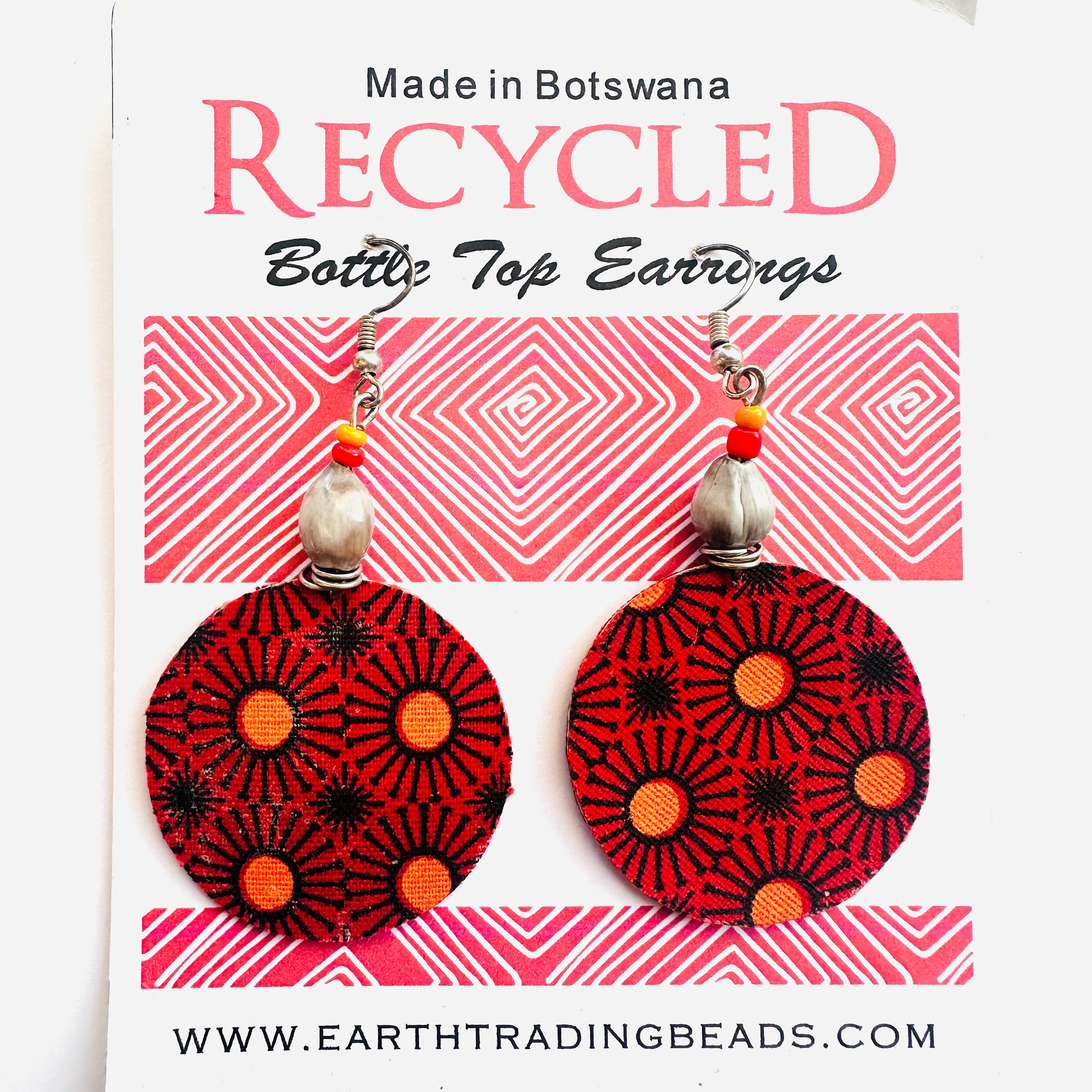 A pair of recycled bottle top earrings with pink, orange, and black geometric patterns, displayed on a card with 'Made in Botswana' text and the brand name 'Earth Trading'.
