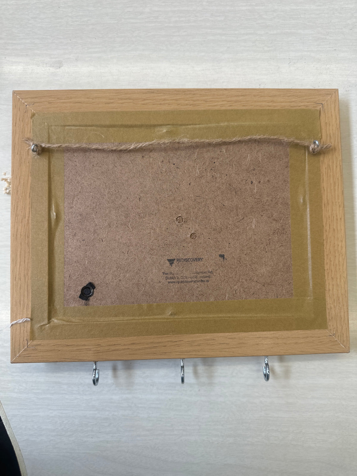 The back view of a square picture frame with a light oak wood border, a brown cardboard backing, and a gold-colored inner liner, shown on a white textured background.