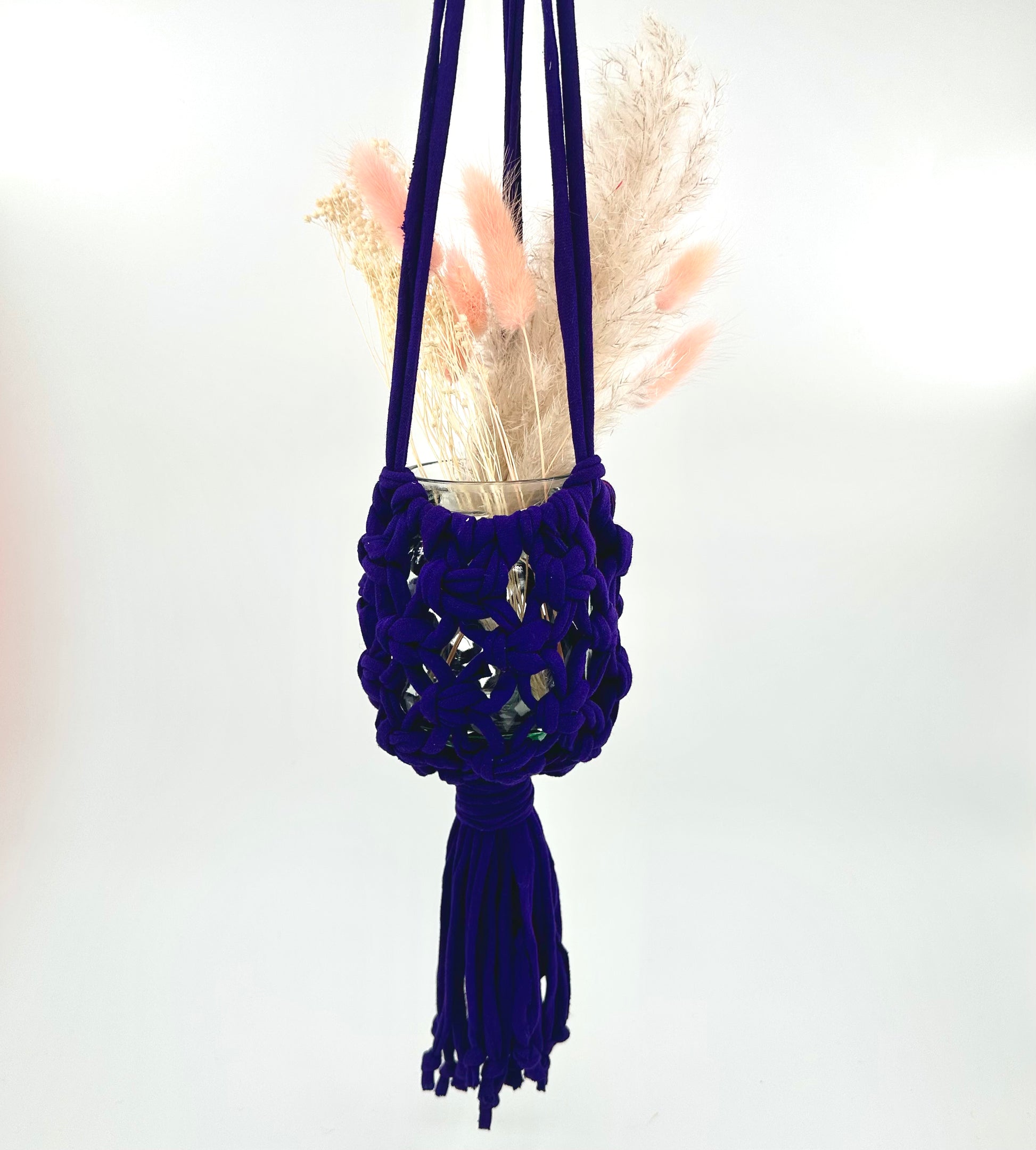 A blue macrame jar holder hanging against a white background, with a tassel at the bottom and containing some decorative elements.
