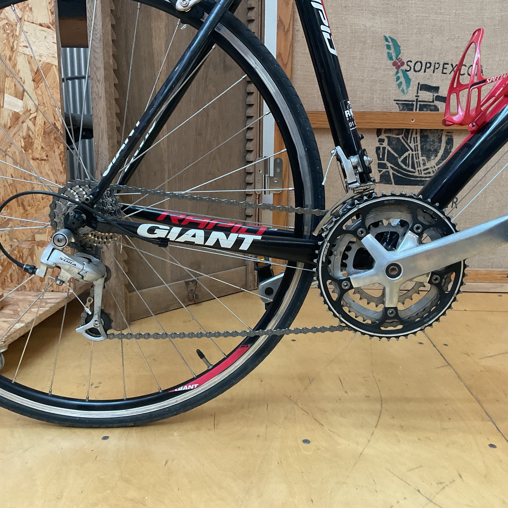 Close-up of a black Giant road bicycle showing the rear wheel, chainring, cassette, derailleur, and part of the frame in a workshop setting.