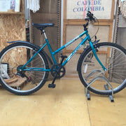 A teal mountain bike with black components and silver rims, displayed on a metal stand in a workshop or shop setting with plywood and a sign in the background.