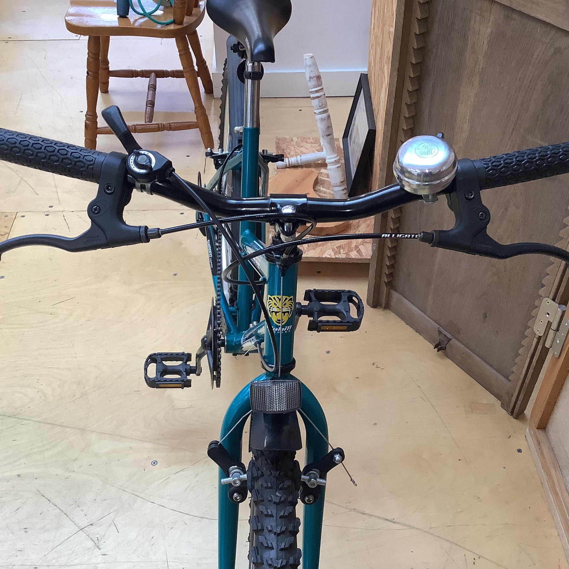 Front view of a teal mountain bike with black handlebars, a silver bell, and black pedals, photographed indoors on a light wood floor.