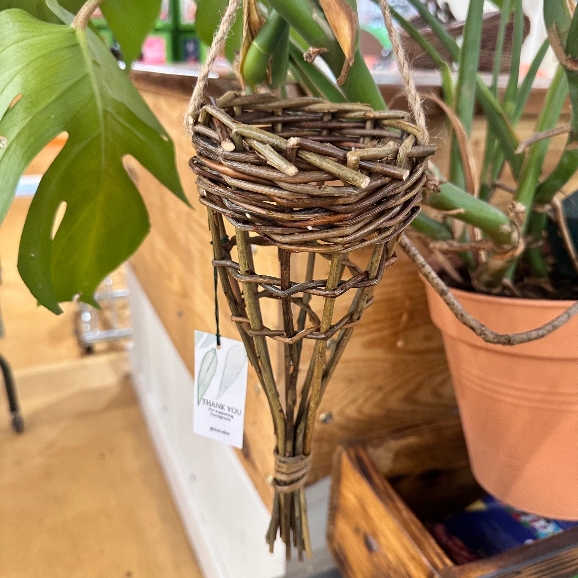 A woven willow bird feeder with visible twine and hanging leaves, displayed in a garden setting.