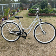 A white step-through city bicycle with black tires, a black saddle, and black chain guard, parked on a gravel path in a backyard garden setting.