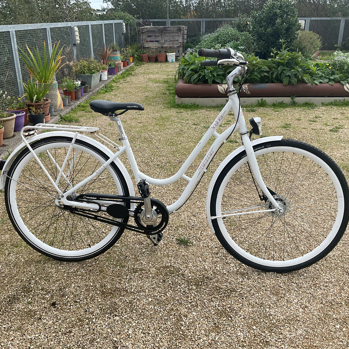 A white step-through city bicycle with black tires, a black saddle, and black chain guard, parked on a gravel path in a backyard garden setting.