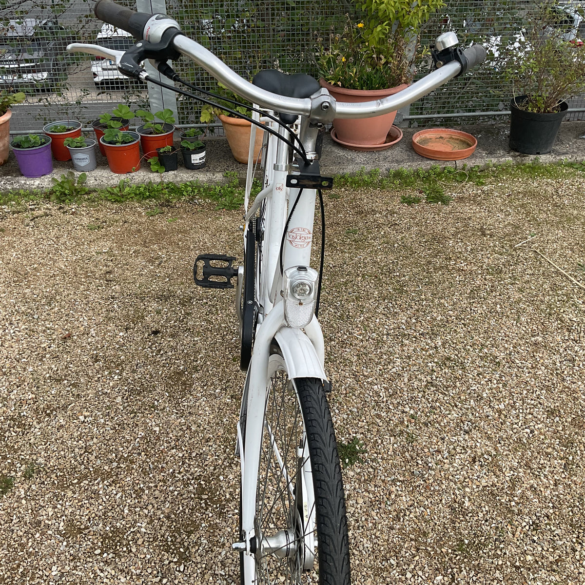 A white step-through bicycle with black tires and silver handlebars, photographed outdoors on a gravel surface with potted plants in the background.