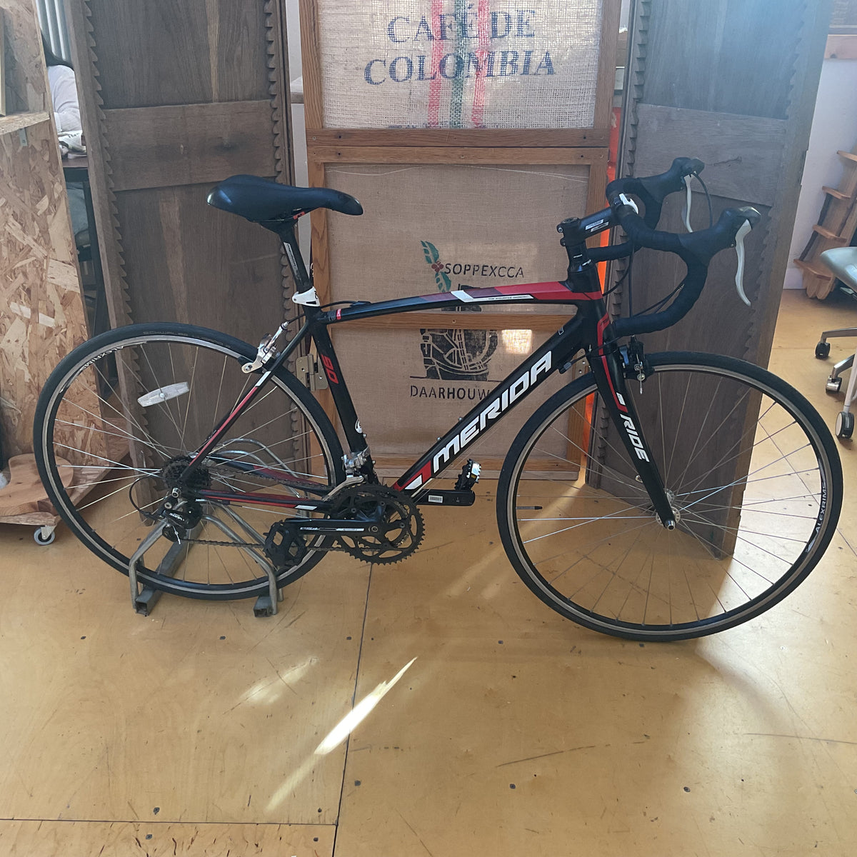 A black and red Merida road bicycle with white branding, displayed indoors on a stand against cardboard crates.