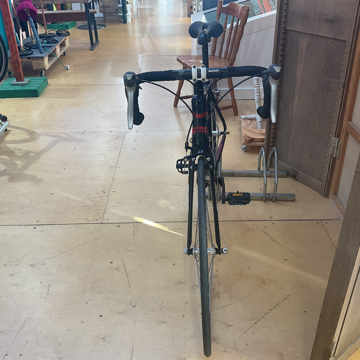 A black drop-bar bicycle photographed from the front inside a shop or showroom with a wood floor and furniture visible in the background.