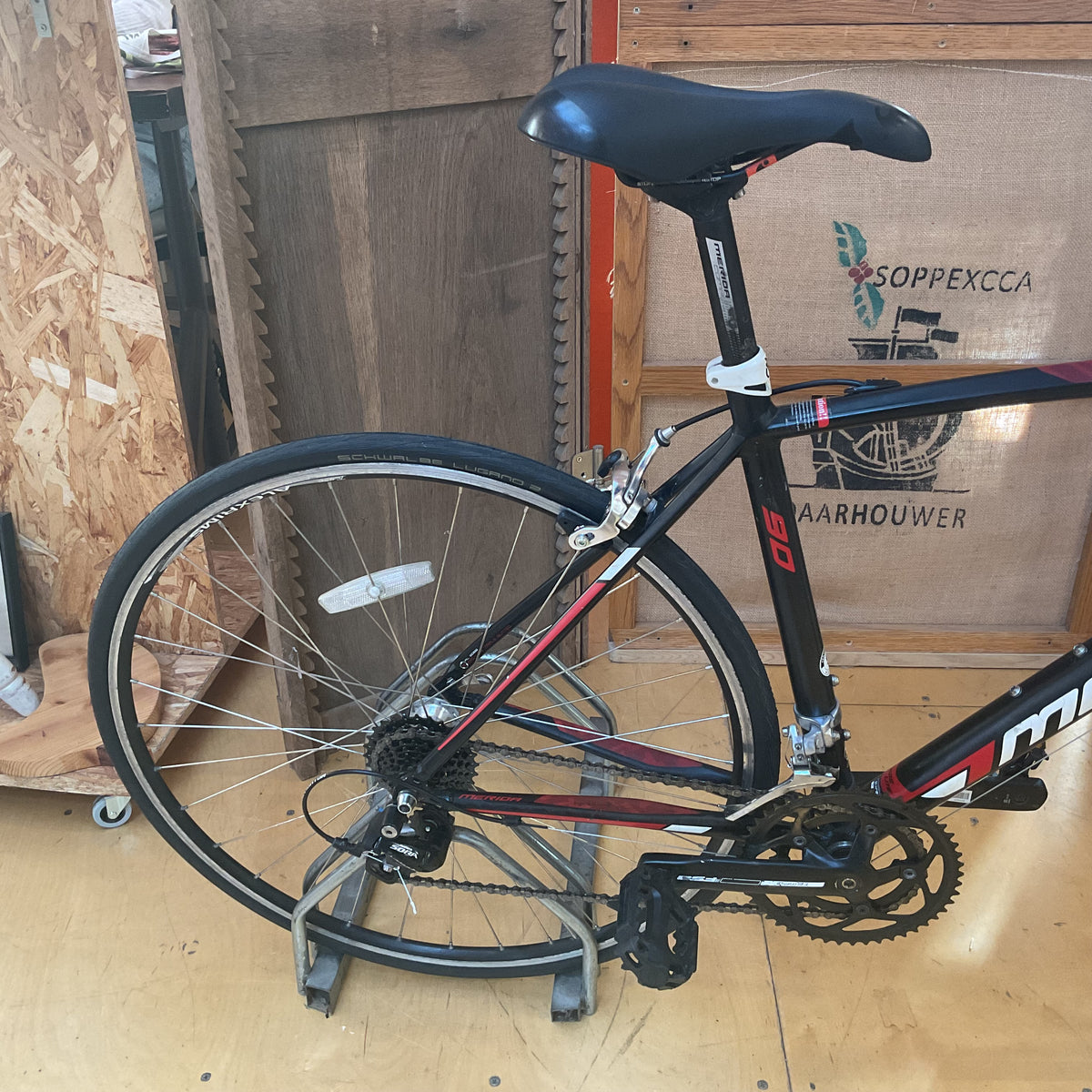 A black mountain bike with red and white graphics, mounted on a metal rack in a workshop. The bike has a black saddle, black handlebars, and a black frame with red and white accents.