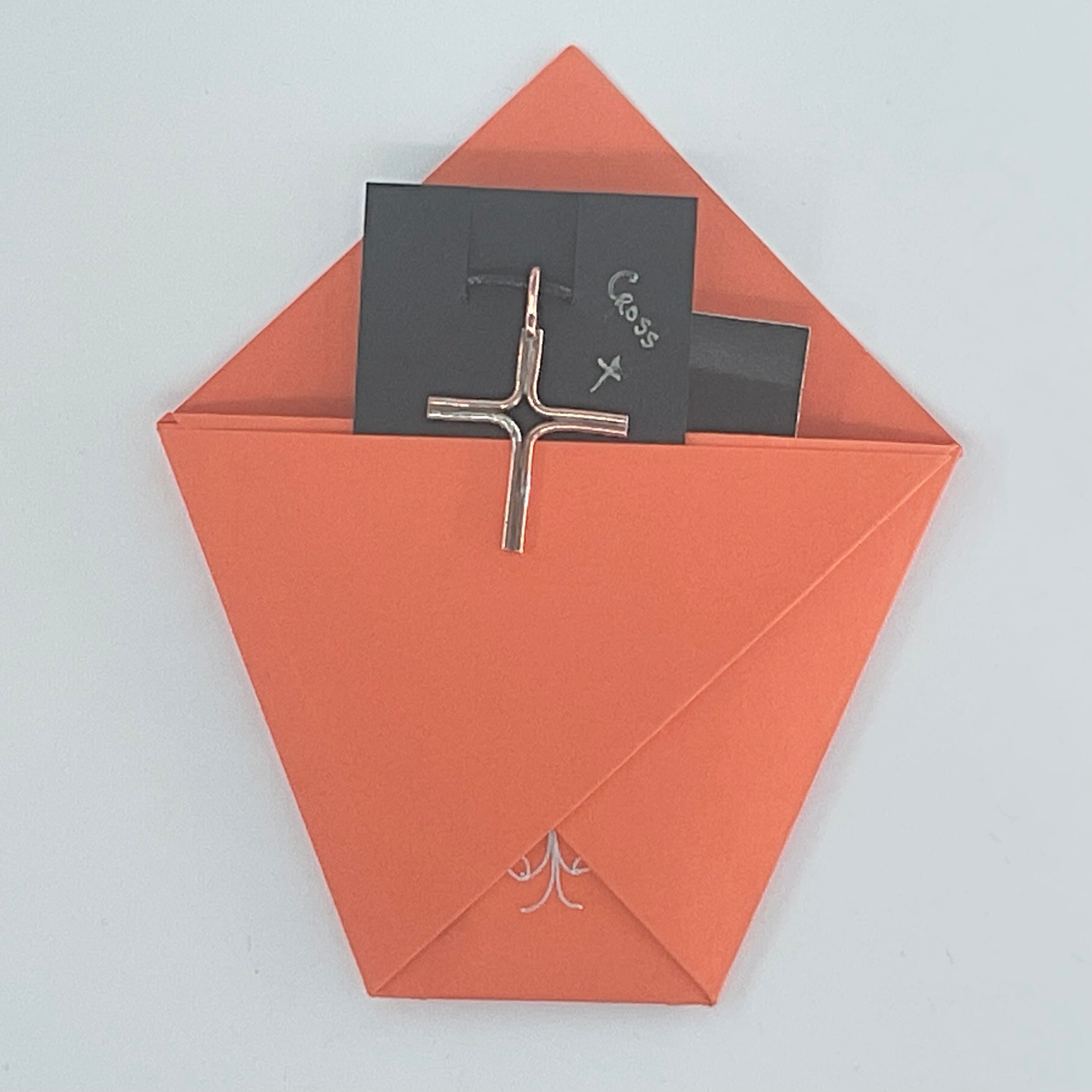 A silver-toned cross pendant with a drop loop, displayed on a black card inside an orange paper envelope against a light gray background.