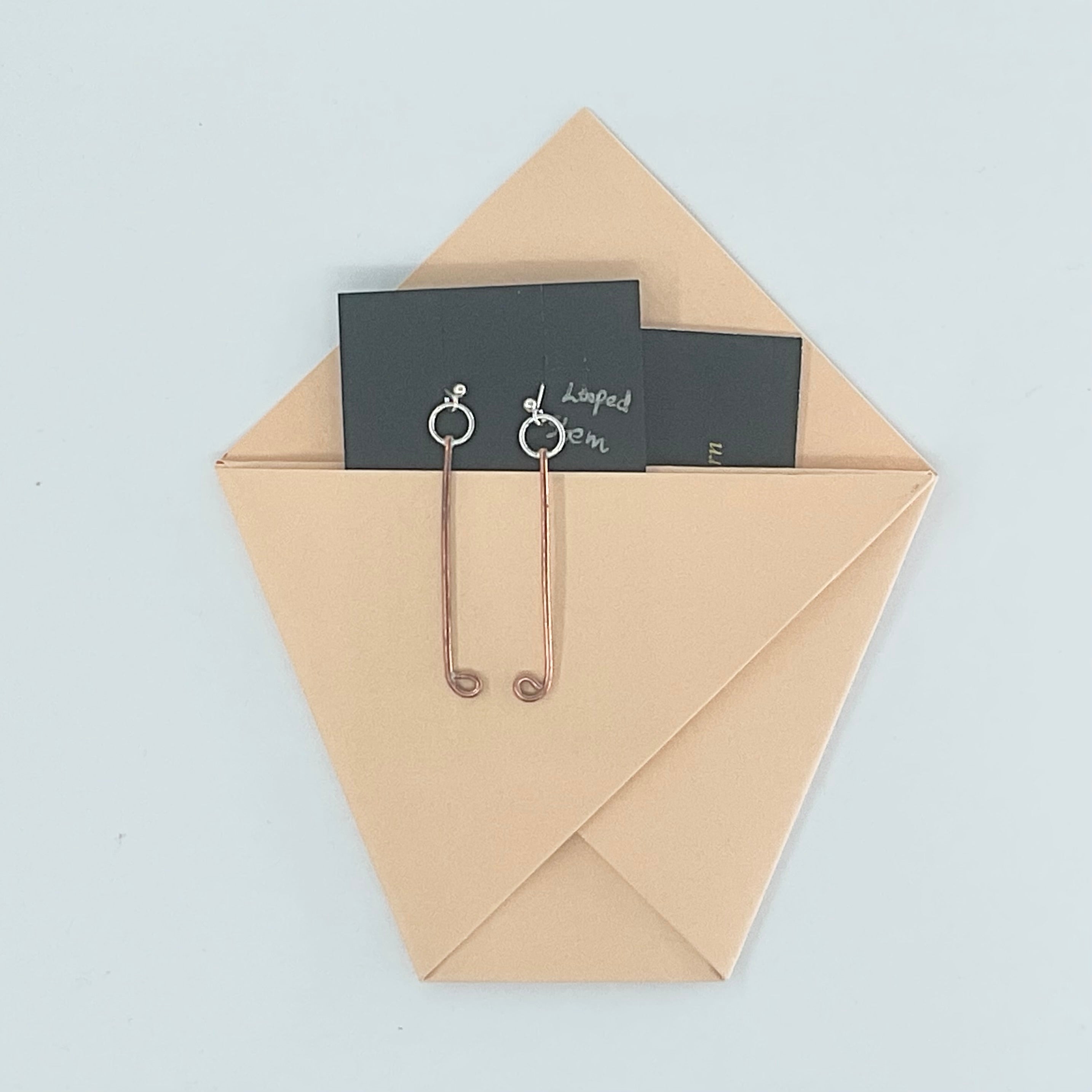A pair of rose gold wire safety pins clipped to black display cards, resting inside a light tan paper envelope on a pale blue background.