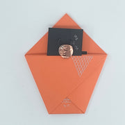 An orange paper envelope or pocket-style card holder with a black card partially pulled out and a round metallic rose-gold coin resting on top, photographed on a plain light background.