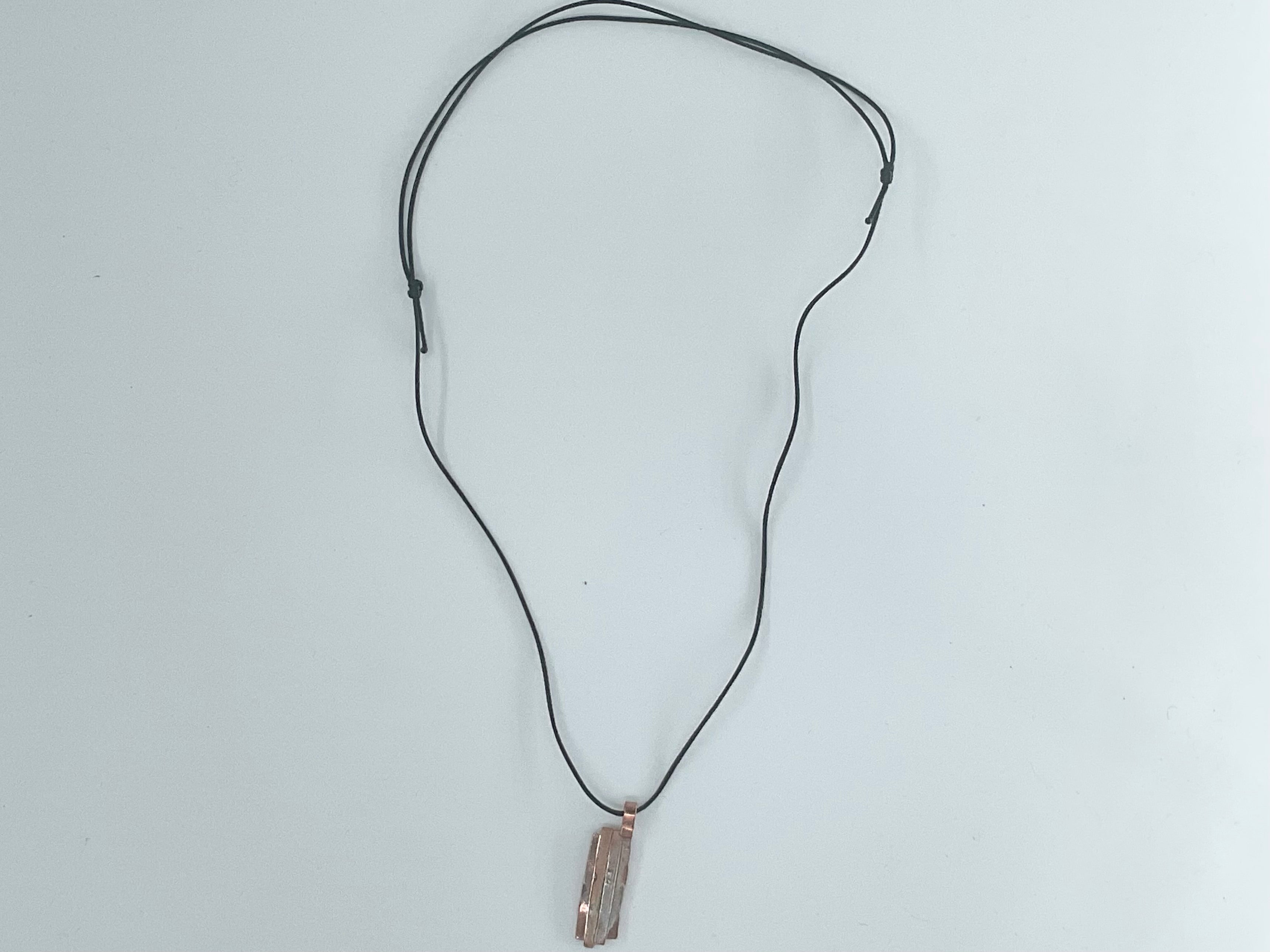 A rose gold rectangular pendant necklace with a clear crystal or glass insert, suspended from a black cord on a light gray background.