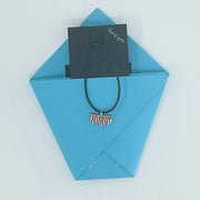 A black cord hair tie with a small metallic barrette-style end, presented on a light blue envelope with a black card labeled 'Bangles' tucked behind it, on a white background.