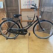A black step-through bicycle with a rear cargo rack, displayed indoors on a wooden floor in front of a rustic wood-paneled wall with a coffee sack sign.