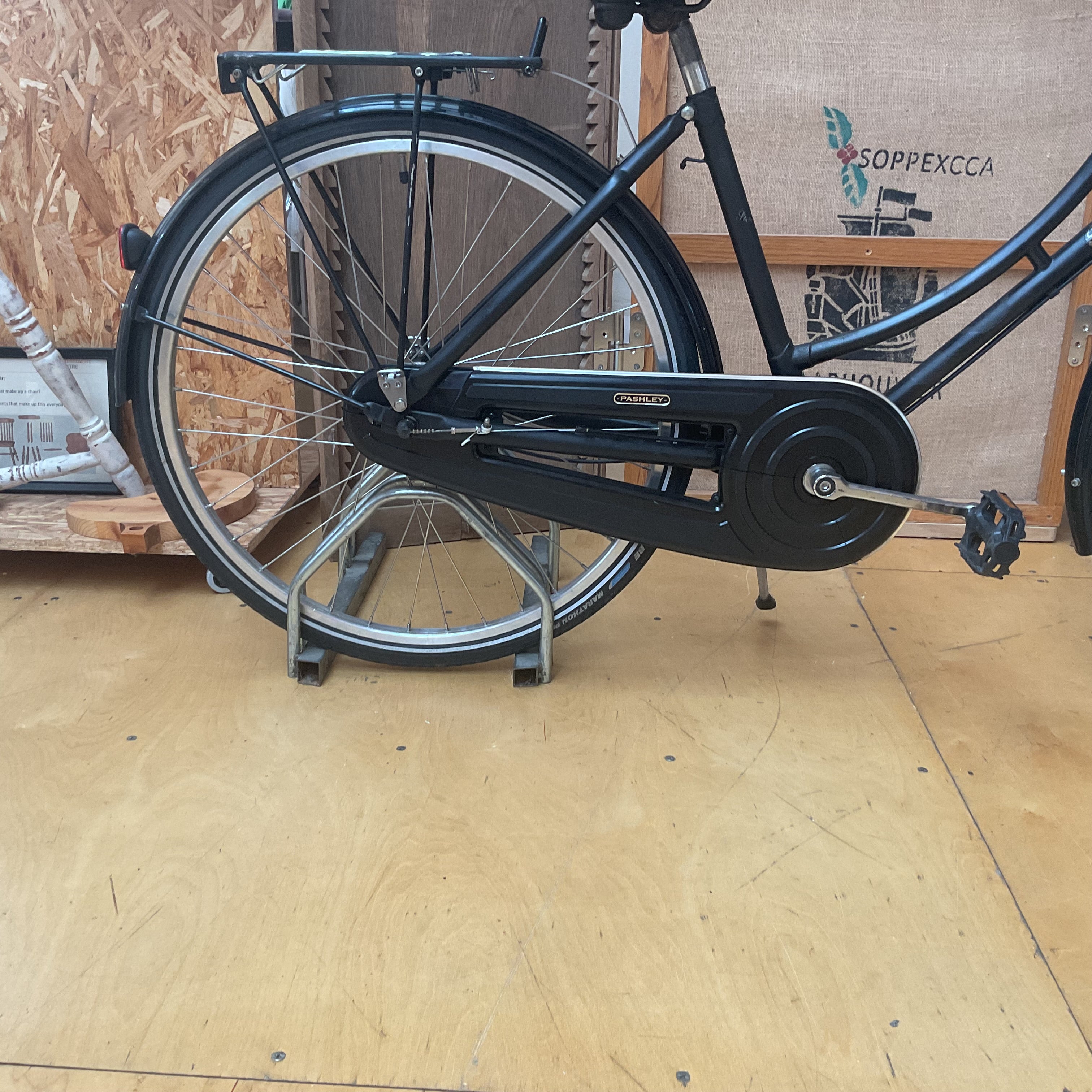 A black Cannond double-decker bicycle with a rear rack and rear wheel elevated on a stand, photographed indoors on a plywood floor against a wood-paneled wall.