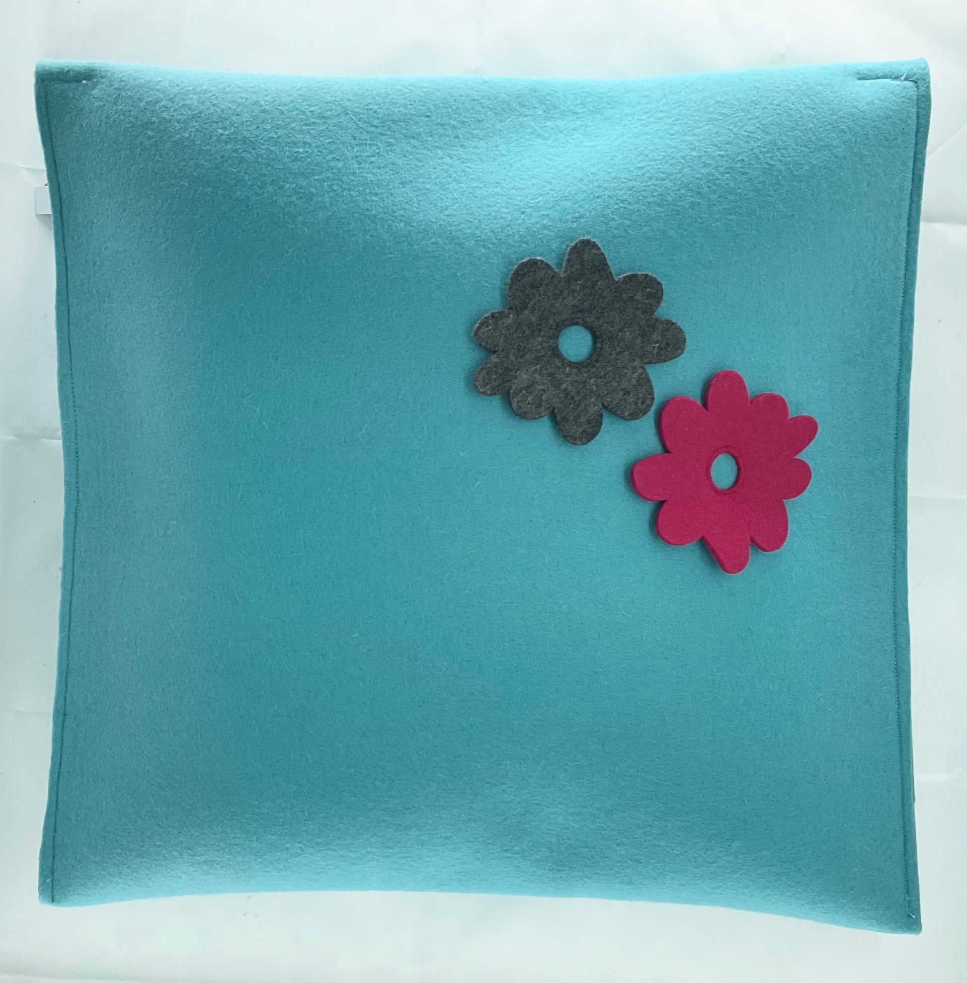 Teal pillow with two felt flowers, one gray and one pink, on a white background