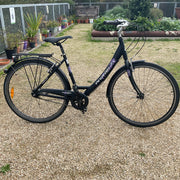 A black step-through bicycle with purple and white 'pedalstreet' branding, parked on a gravel path in a garden setting.