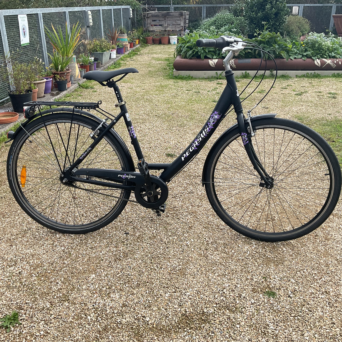 A black step-through bicycle with purple and white 'pedalstreet' branding, parked on a gravel path in a garden setting.