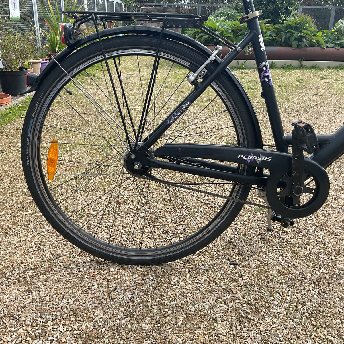 Close-up of the rear wheel and frame of a black Pelicanus folding bicycle parked on a gravel surface outdoors.