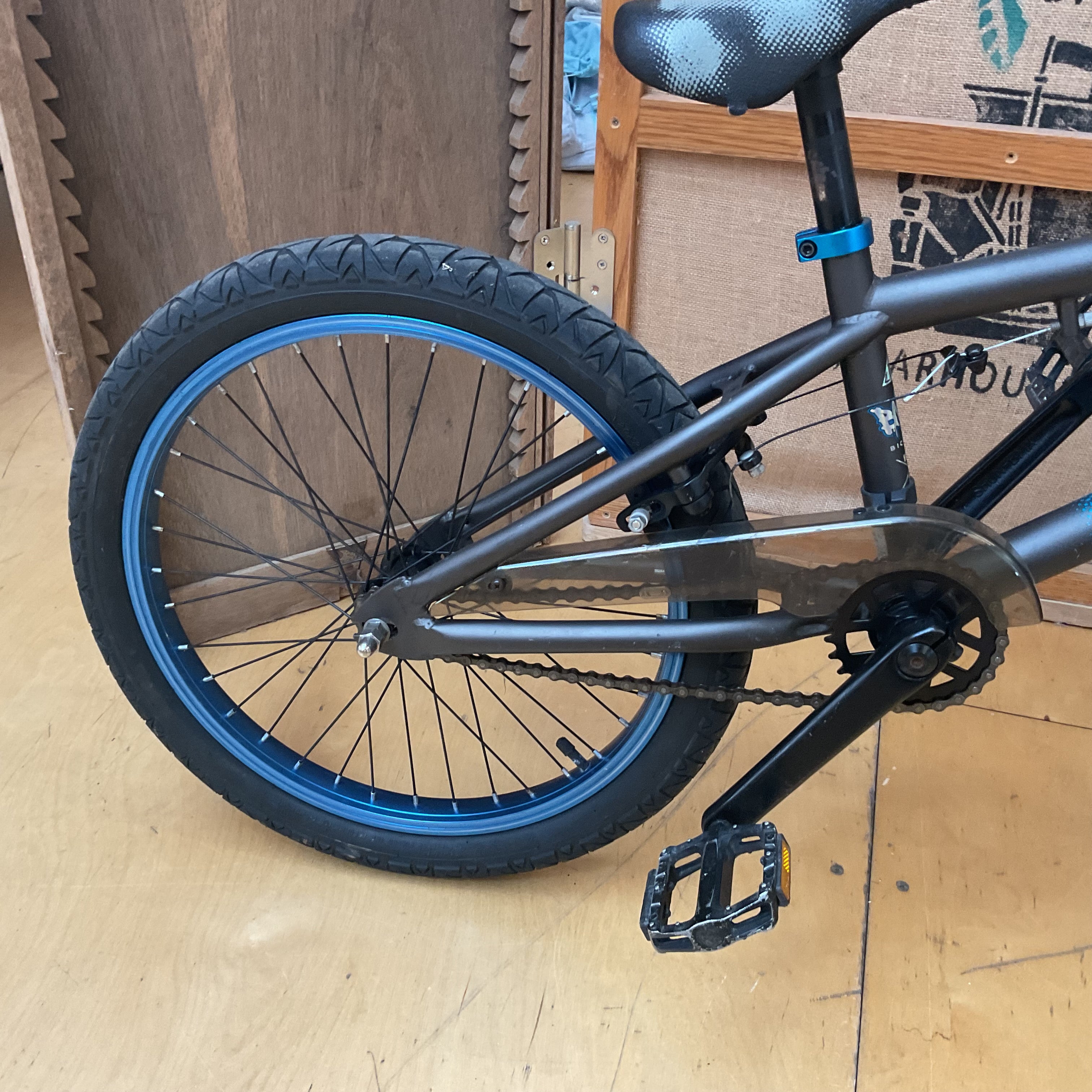 Close-up of the rear wheel and frame of a BMX bike with a matte black frame, black tire, and bright blue rim accents, photographed indoors on a wooden floor.