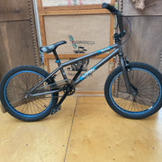 A dark gray BMX bike with blue accents on the rims and frame details, displayed on a wooden floor in front of a wooden wall and a burlap sign.