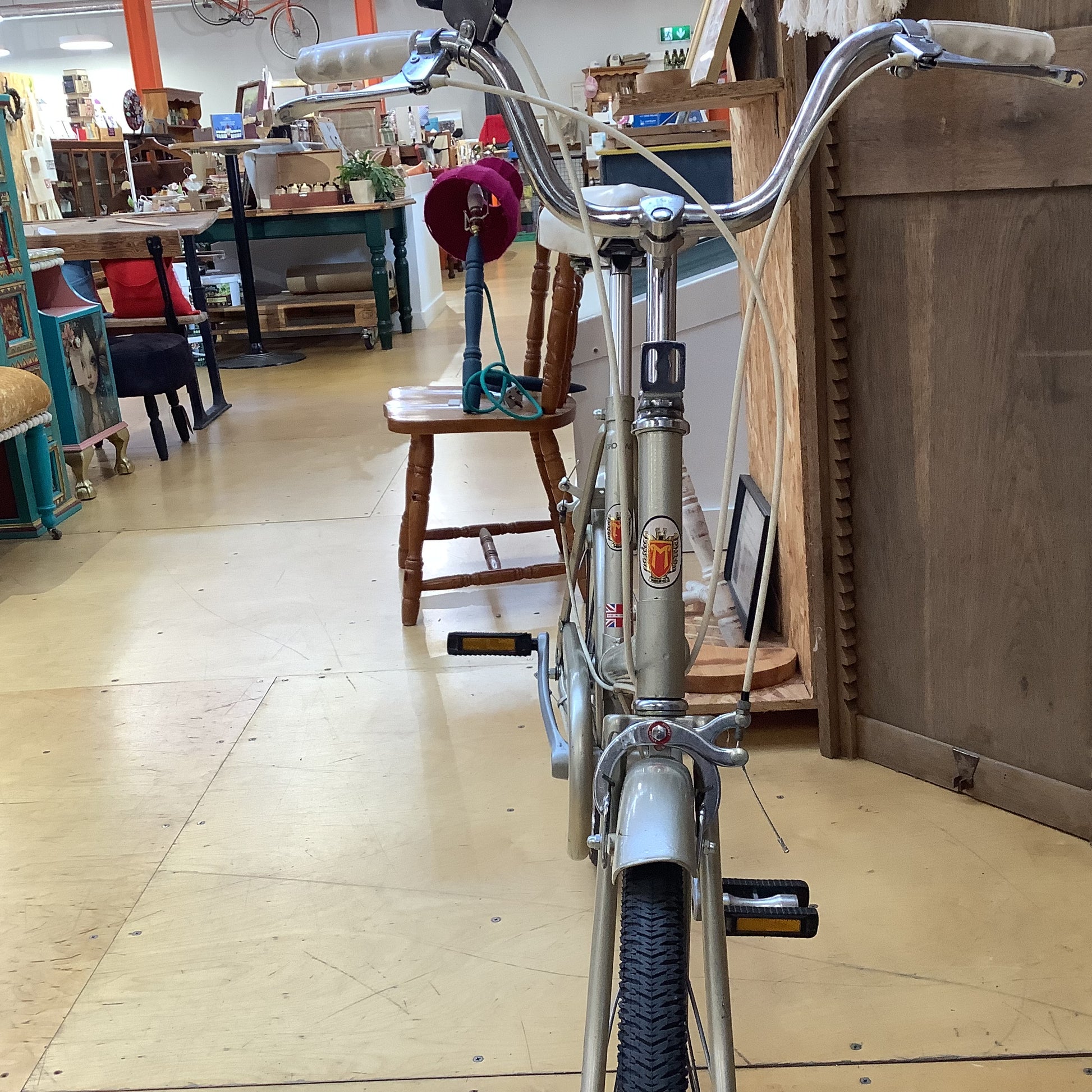 A vintage silver bicycle with high handlebars and a step-through frame, photographed inside a cluttered antique shop or furniture store.