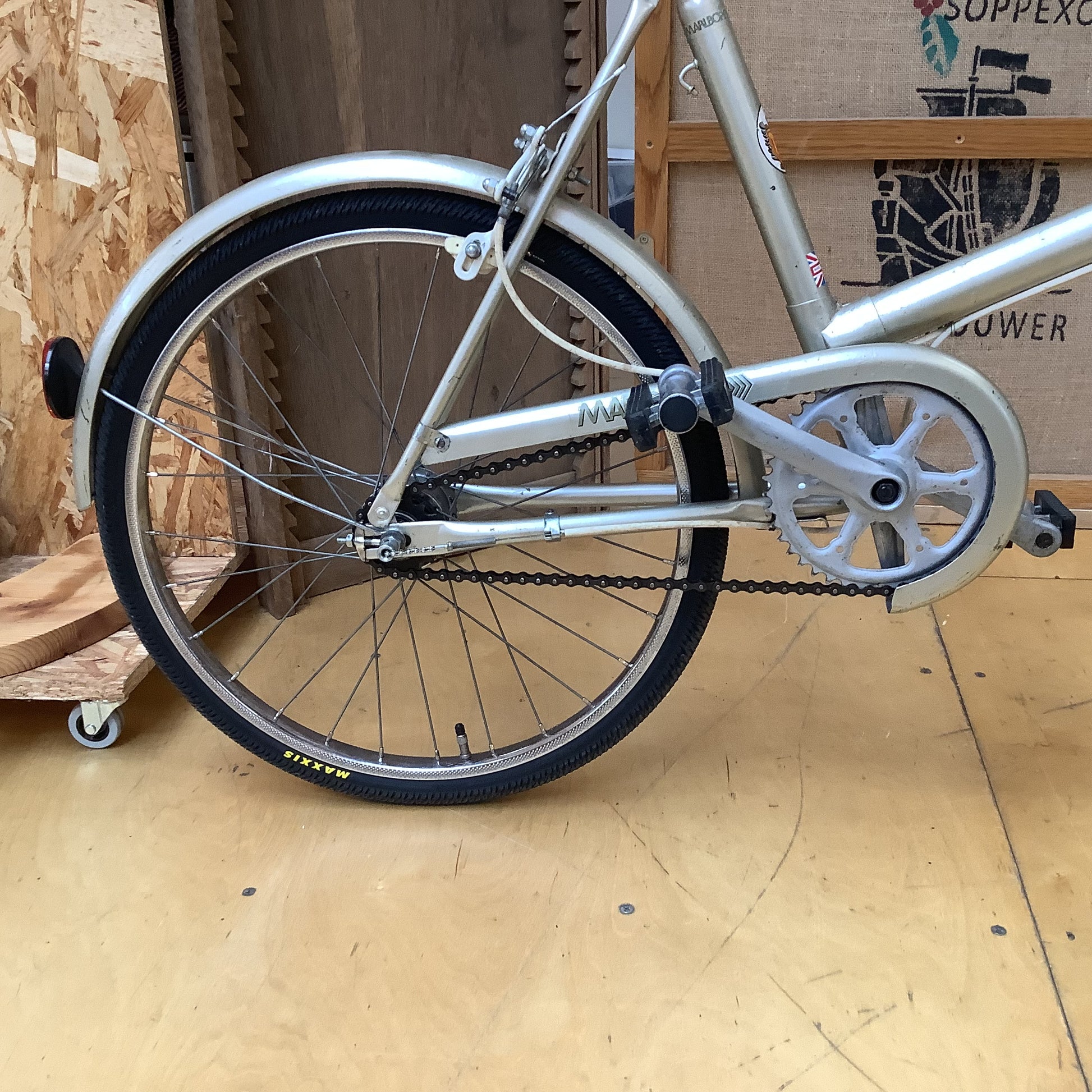 A close-up side view of a vintage silver bicycle frame, rear wheel, chain guard, and chainstay, set against a workshop background with plywood and a cardboard box.