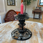 A black bicycle disc and chainring candle holder with a pink candle on top, displayed on a rustic wooden table in a vintage-style room.