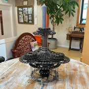 A handmade metal sculpture centerpiece made from stacked sprockets, gears, and chains, holding a tall gray candle, displayed on a wooden table in a shop interior.