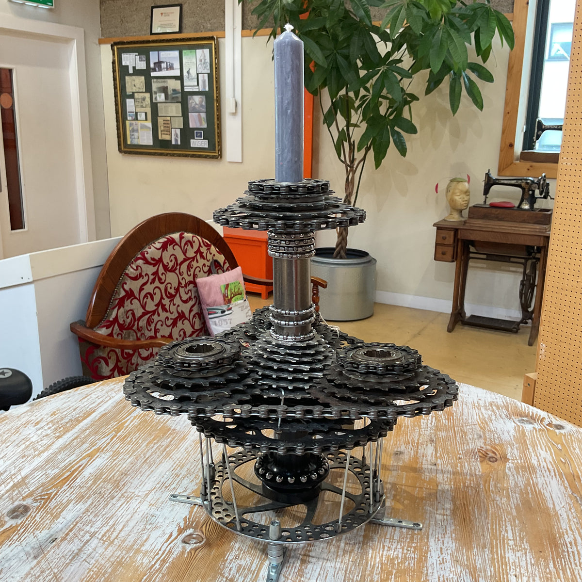 A handmade metal sculpture centerpiece made from stacked sprockets, gears, and chains, holding a tall gray candle, displayed on a wooden table in a shop interior.