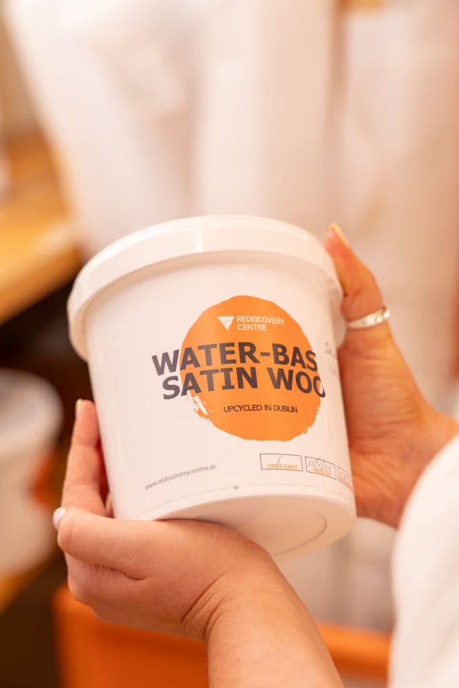 A person holds a white tub of Rediscovery Centre WaterBased Satin Wood paint in a warm, softly blurred indoor setting. The tub features a prominent orange circular label with black text.