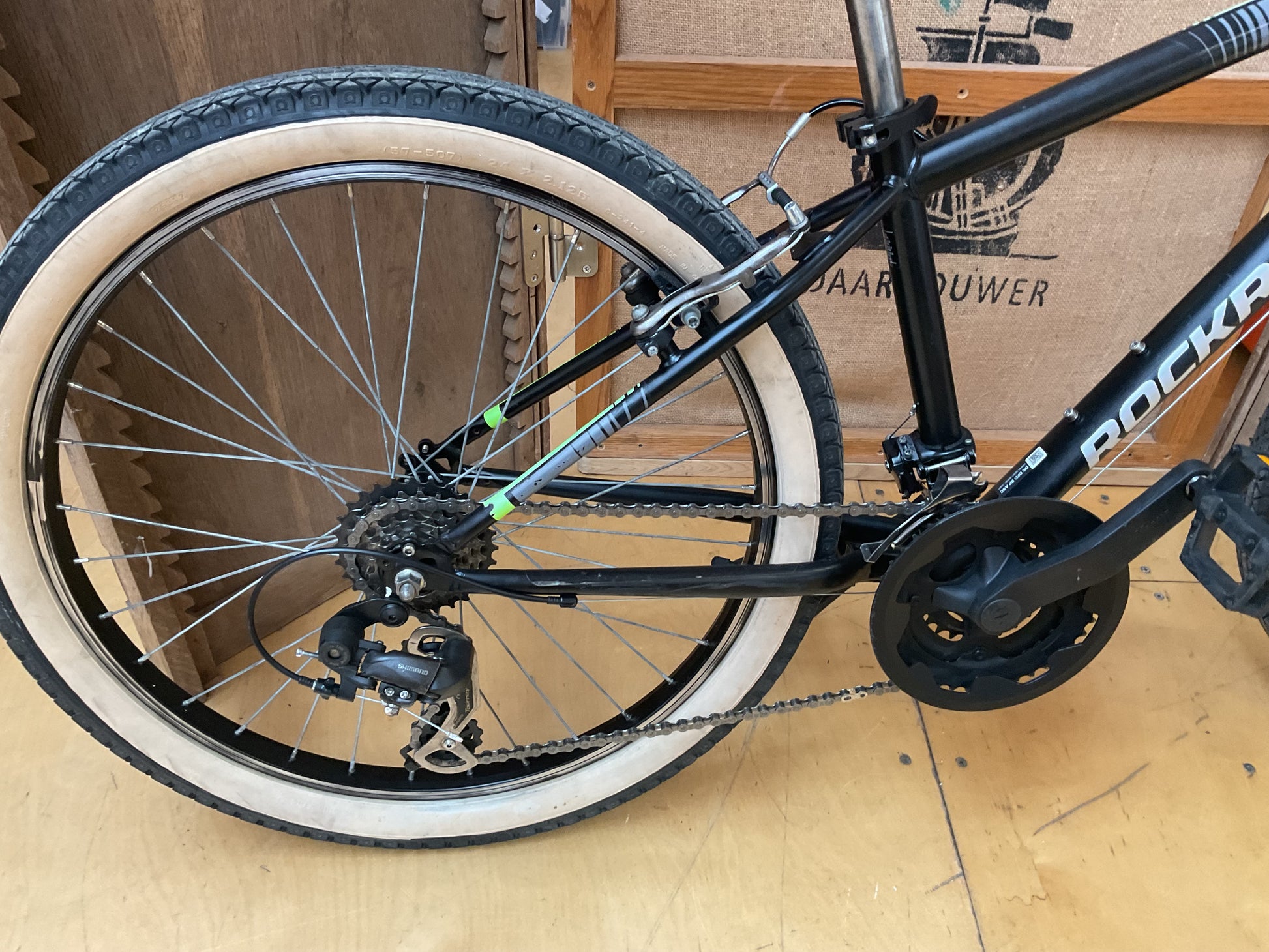 Close-up of a black Rockhopper bicycle frame, rear wheel, chainring, and rear derailleur, photographed indoors against a cardboard box and wood floor.