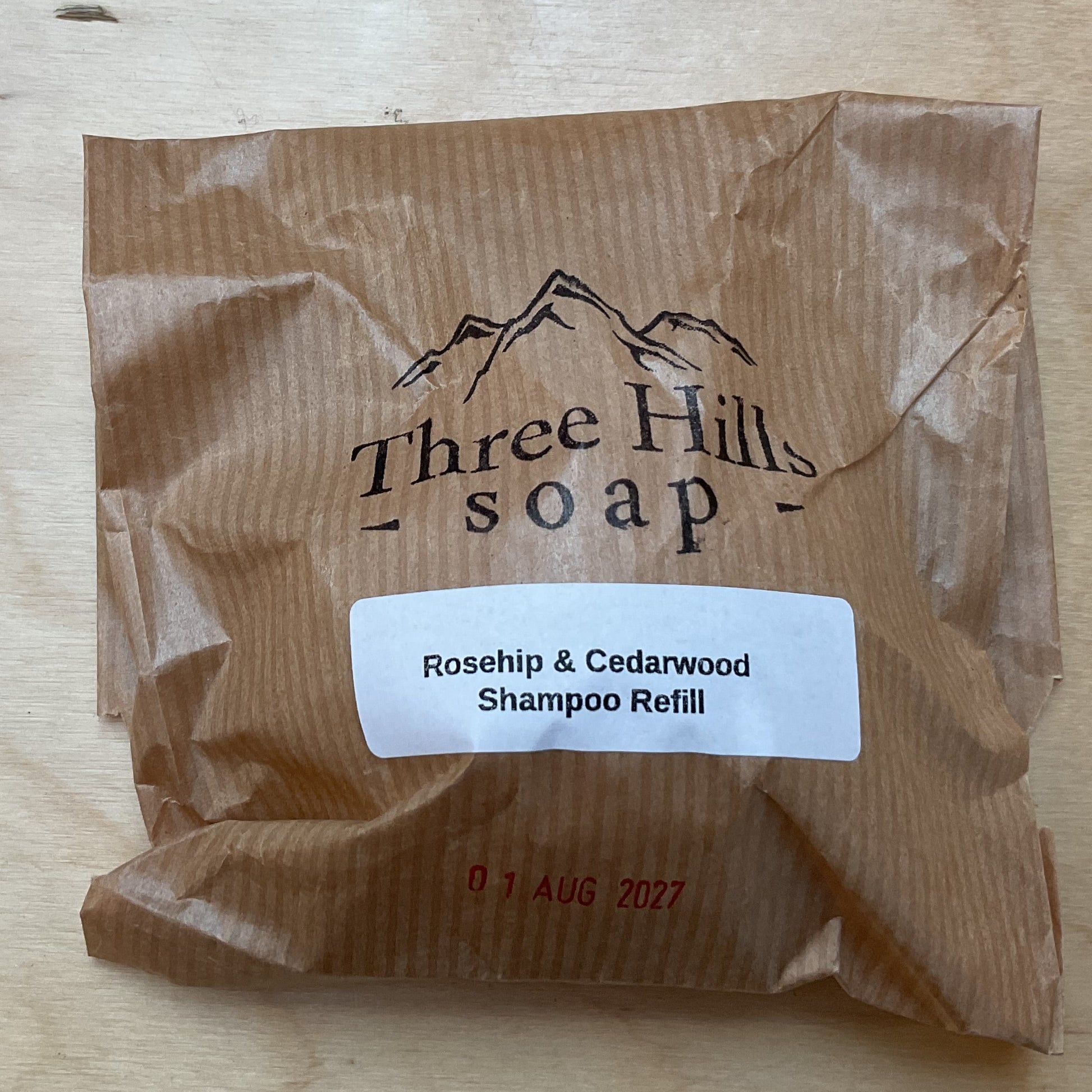 A brown kraft paper refill pouch for Three Hills Soap, labeled 'Rosehip & Cedarwood Shampoo Refill', placed on a light wood grain surface.