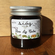 A jar of Sióg of the Earth Rose Lip Balm with a label showing the brand name, product name, and ingredients such as rose oil, almond oil, beeswax, honey, vitamin E, and aloe vera gel.
