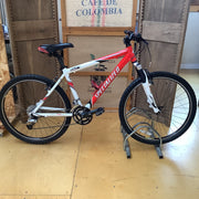 A Specialized mountain bike with a red and white frame, displayed on a metal stand in front of wooden crates and a burlap sack in a warehouse-like setting.