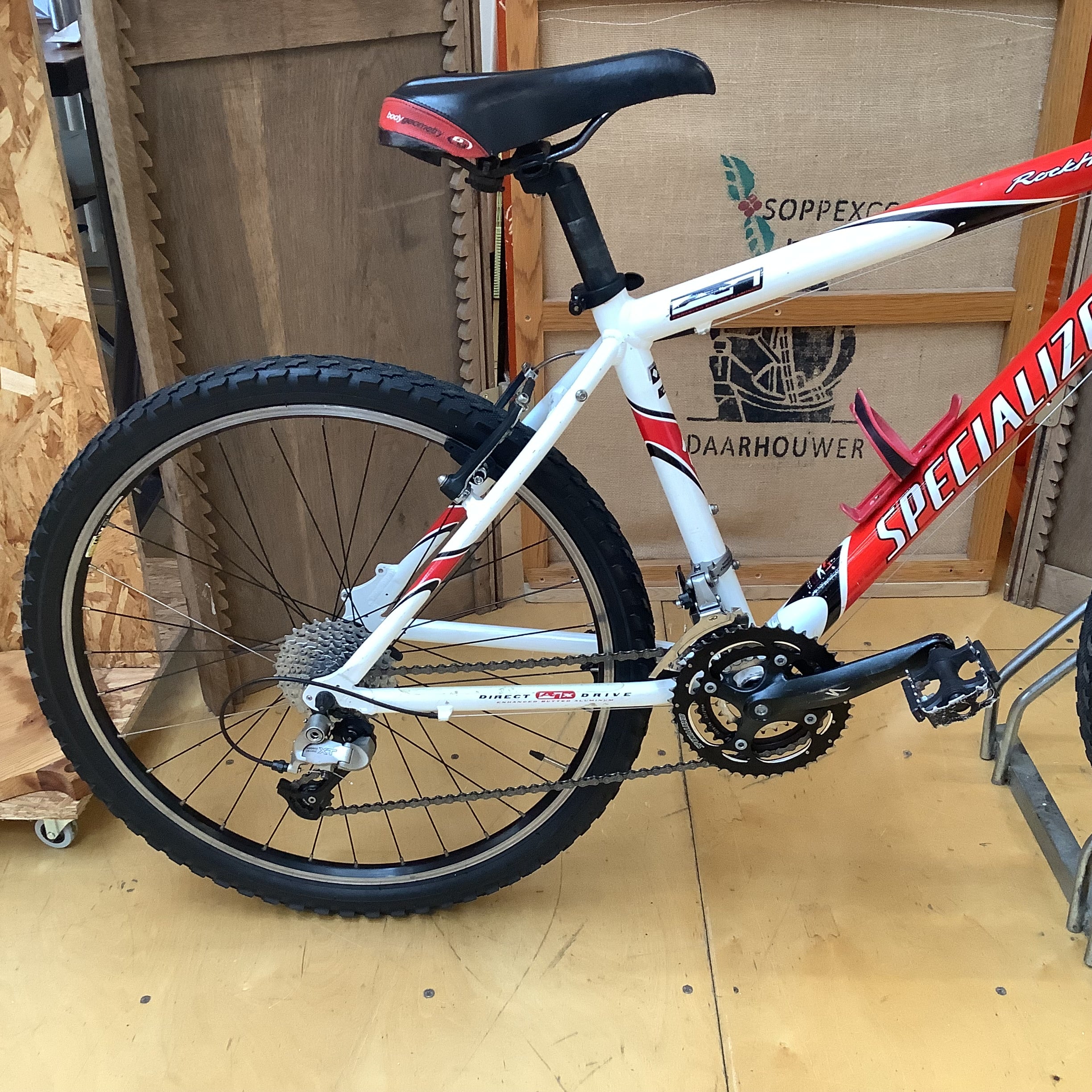 A Specialized road bike with a white frame and red accents, photographed indoors on a wooden floor against a plywood backdrop.