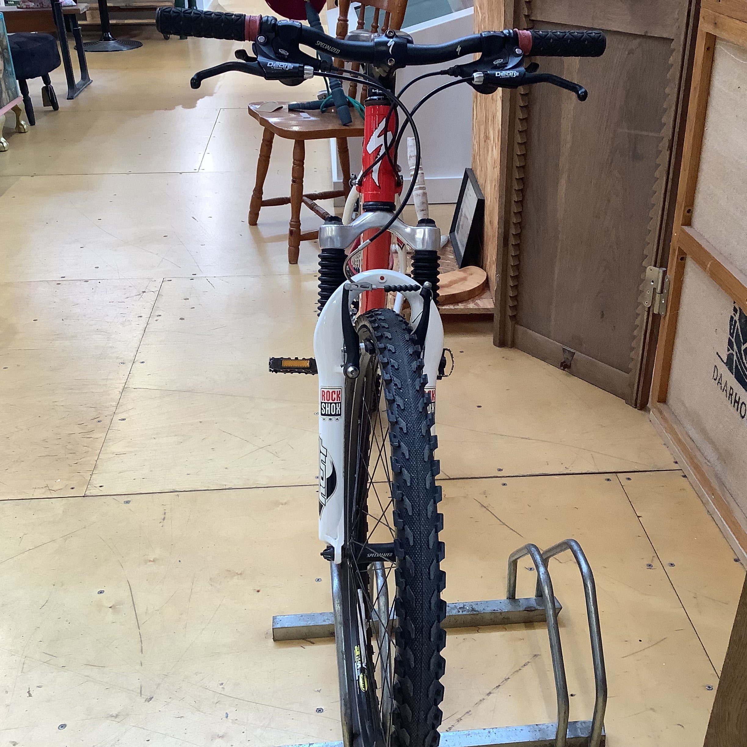 A red and white mountain bike with knobby tires, parked on a metal rack inside a shop with wooden floors and doors.