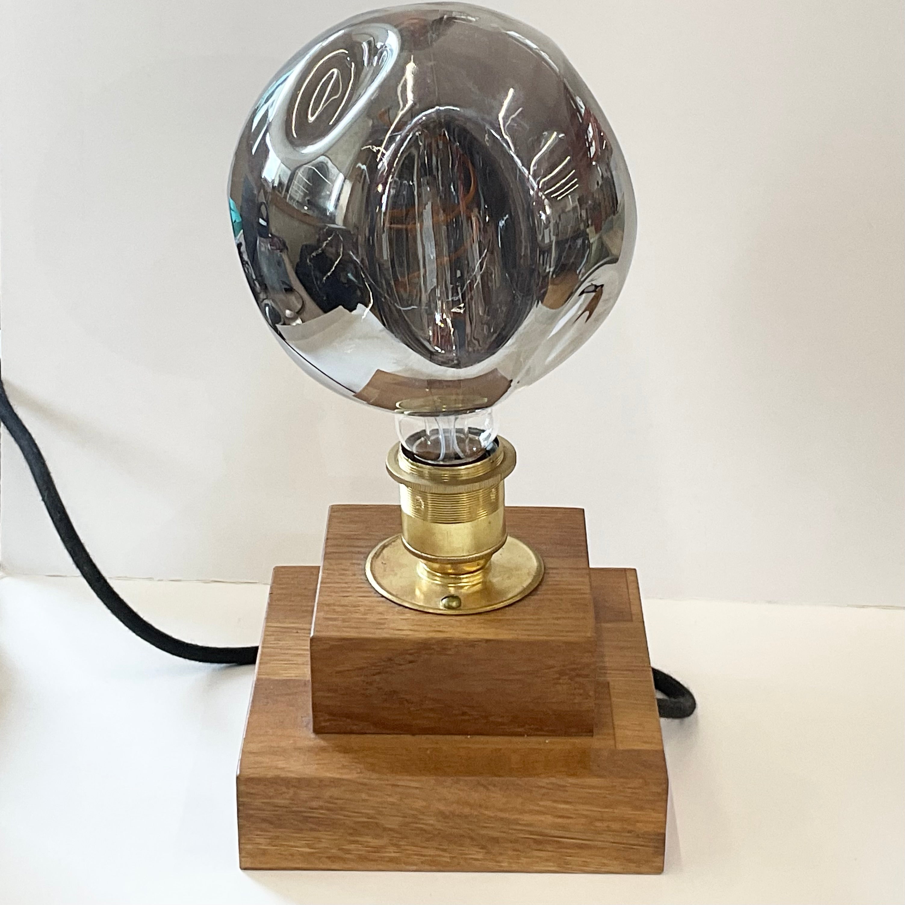 A modern table lamp featuring a large, reflective clear glass globe shade on a polished brass socket, mounted on a two-tiered wooden base with a black power cord.